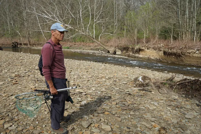 A man in a baseball cap holds fishing equipment while standing at the edge of a stream.