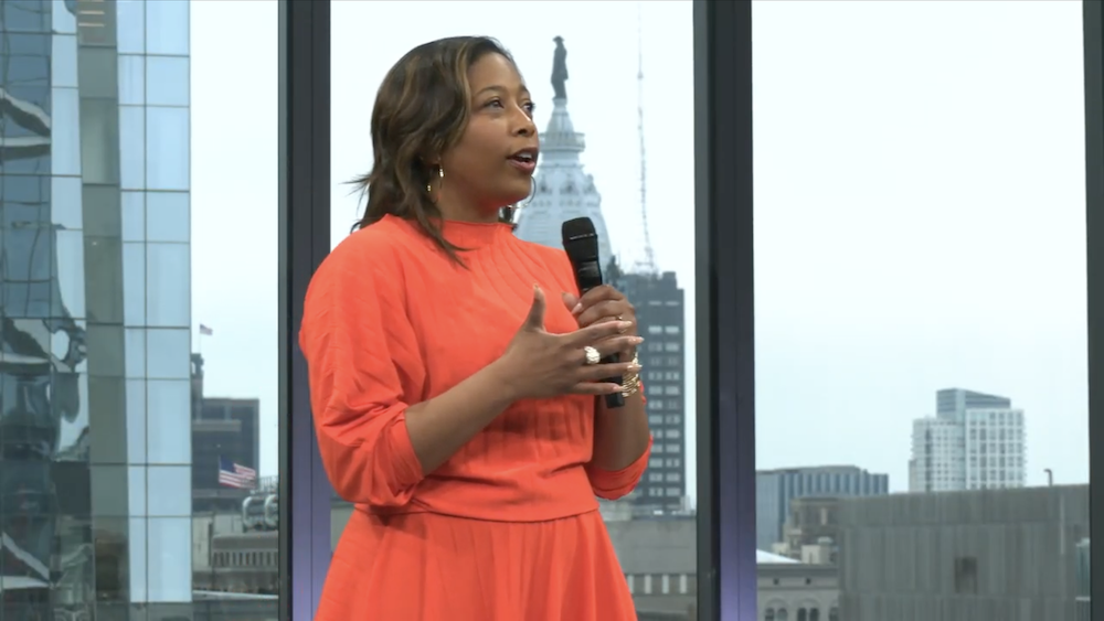 A woman in an orange dress speaks into a microphone with city buildings visible through large windows behind her.