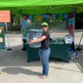 A woman in a neon green baseball hat holding a large gray bucket, stands under a neon green tent with a table behind her.