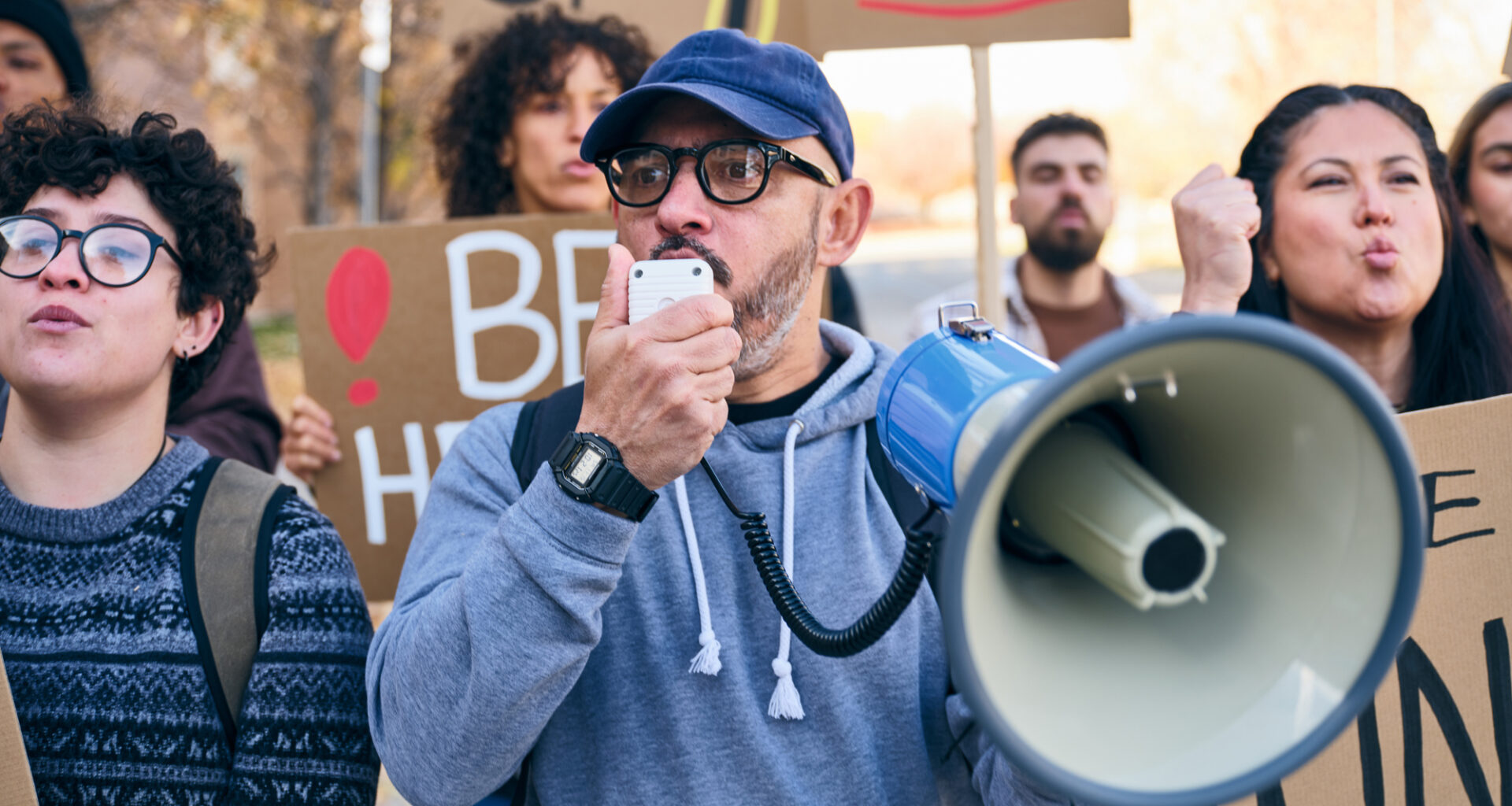 A diverse group of adults gathered outdoors holding handmade signs while a man speaks through a megaphone during a positive community rally.