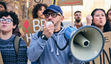 A diverse group of adults gathered outdoors holding handmade signs while a man speaks through a megaphone during a positive community rally.