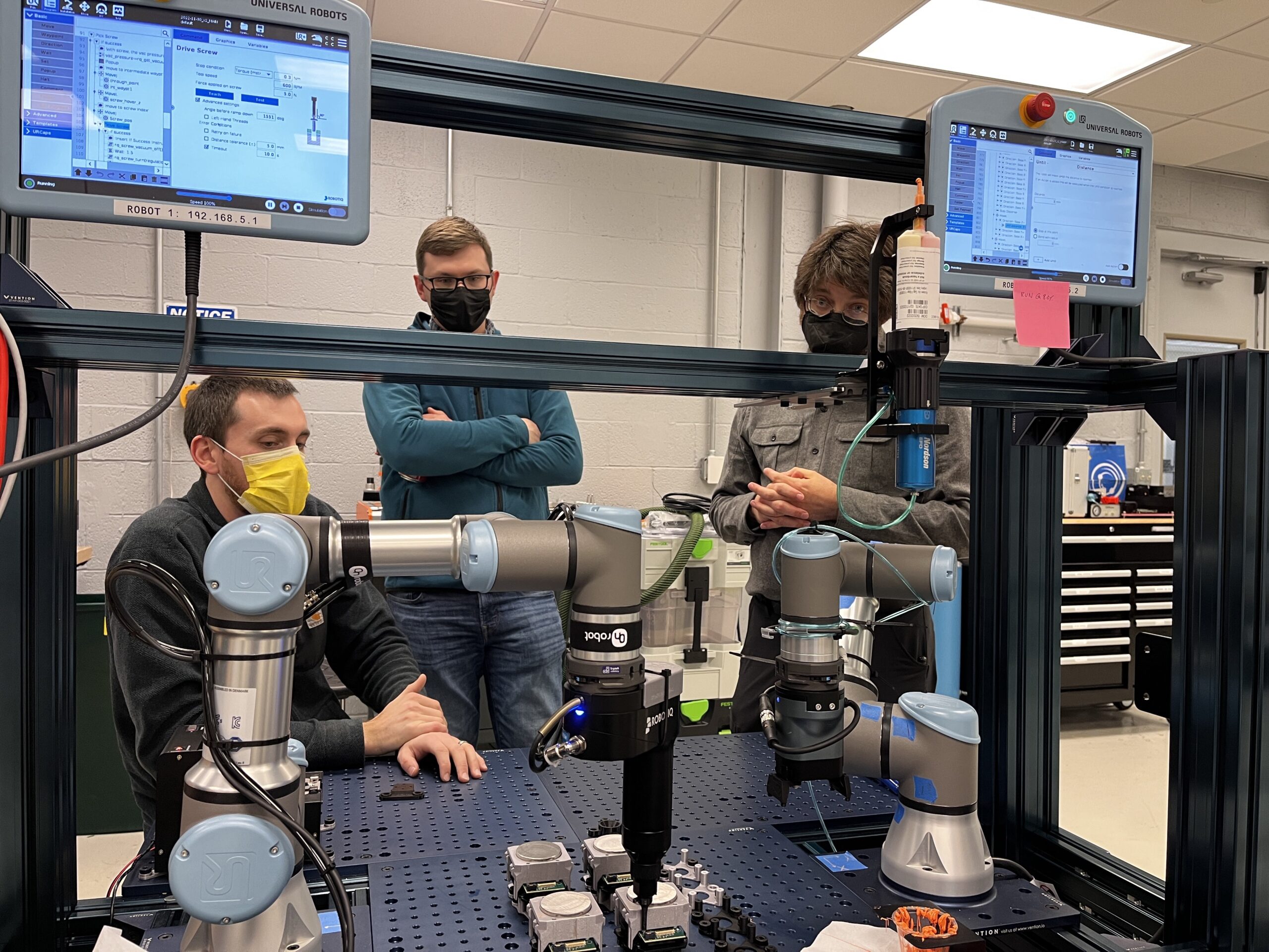 Three people with masks observe and operate a robotic arm workstation in a lab setting, with monitors displaying data and controls.