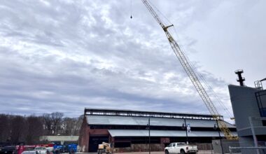 a construction crane rises above the brick turn & grind shop building at steelstacks in bethlehem, with a partially installed metal roof, work vehicles and fencing in the foreground under an overcast sky
