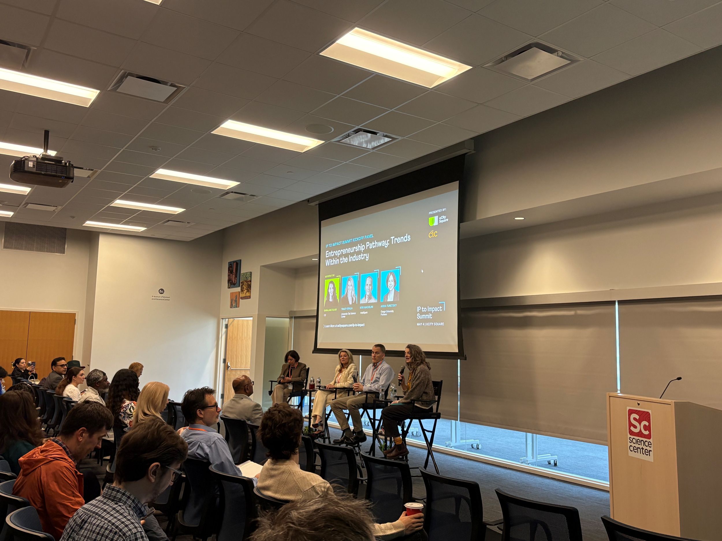 A panel of four speakers sits on stage discussing AI and entrepreneurship trends, with an audience seated in front and a presentation projected on a screen behind them.