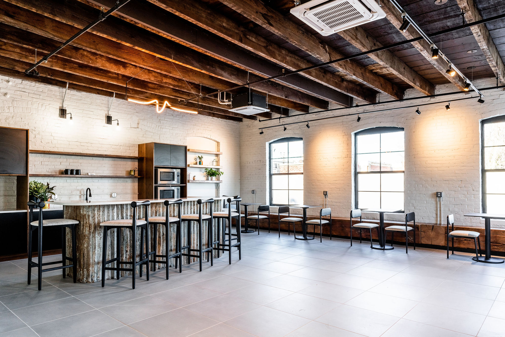 Modern industrial-style kitchen and lounge area with wood and metal finishes, bar seating, large windows, exposed beams, and minimalist decor.