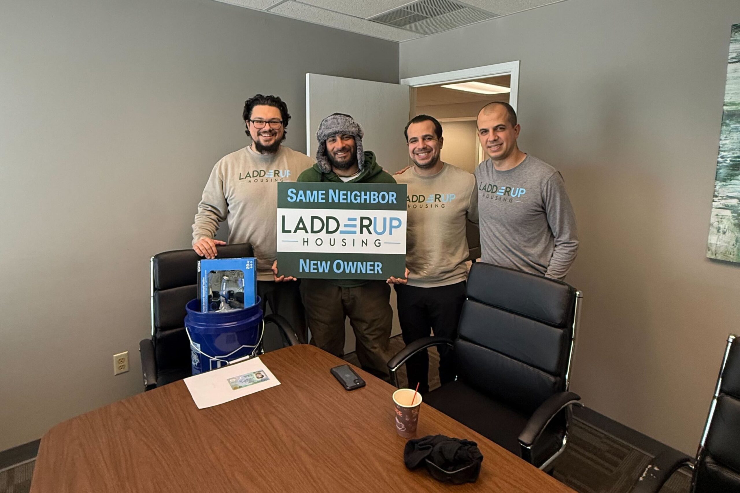 Four men stand in an office, three wearing “Ladder Up Housing” shirts. They hold a sign reading “Same Neighbor, Ladder Up Housing, New Owner” and smile at the camera.