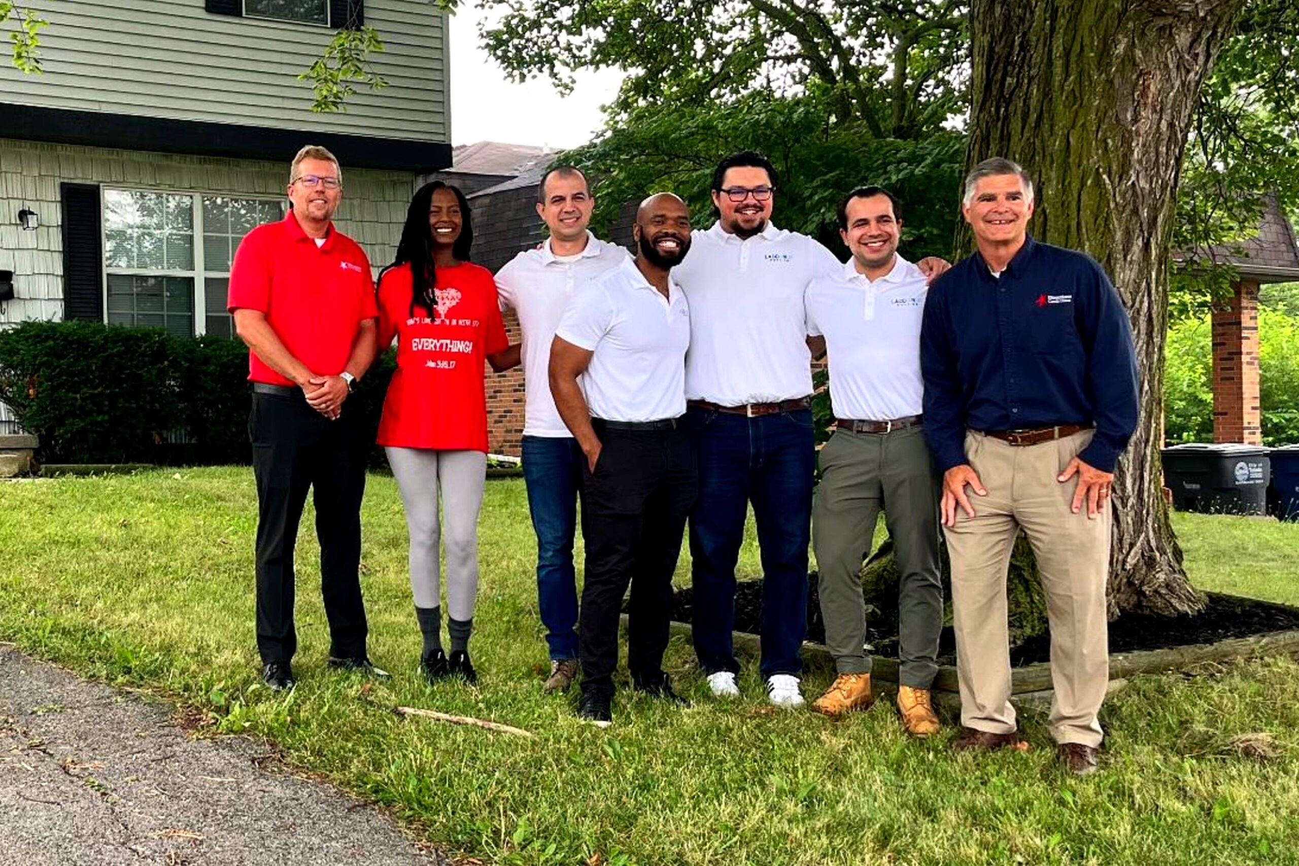 Six people standing and smiling in a row on grass outside a house, with trees and a sidewalk in the background.