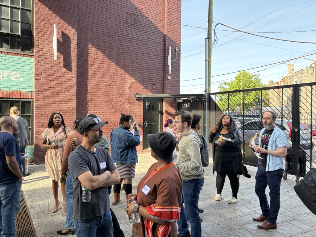 A group of people stand and talk outside near a brick building and metal gate on a sunny day; some hold drinks and wear name tags.
