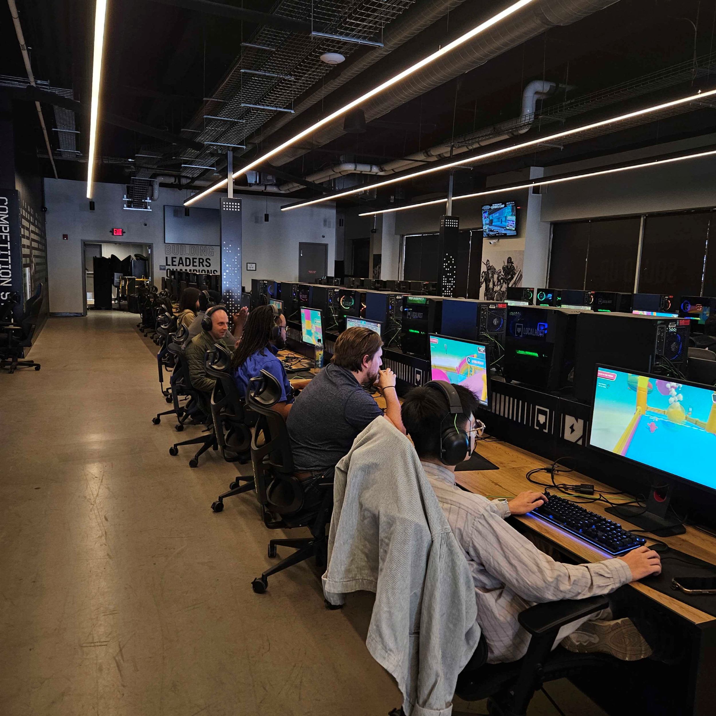 Several people sit at desks with computers in a gaming room, each focused on monitors displaying a colorful video game.