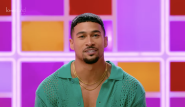 Sean Reifel, a Bethlehem police officer cast on "Love Island USA" season 8, smiles in an open green knit polo shirt and two gold chains, against a backlit pink, purple, orange and red grid wall with the "Love Island USA" logo in the upper left.