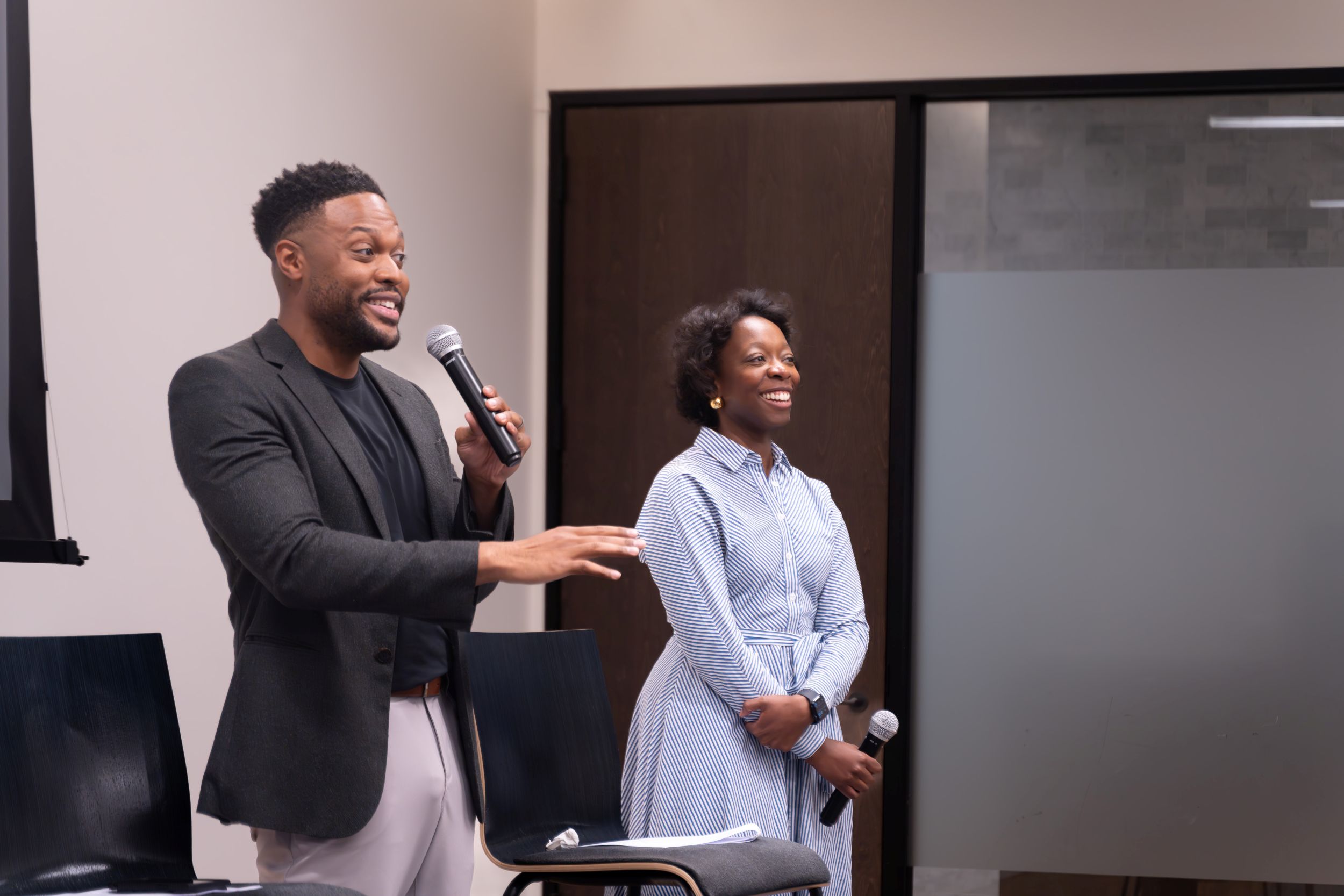A man speaks into a microphone while gesturing, and a woman stands beside him smiling, also holding a microphone. They are indoors next to a chair and a frosted glass door.