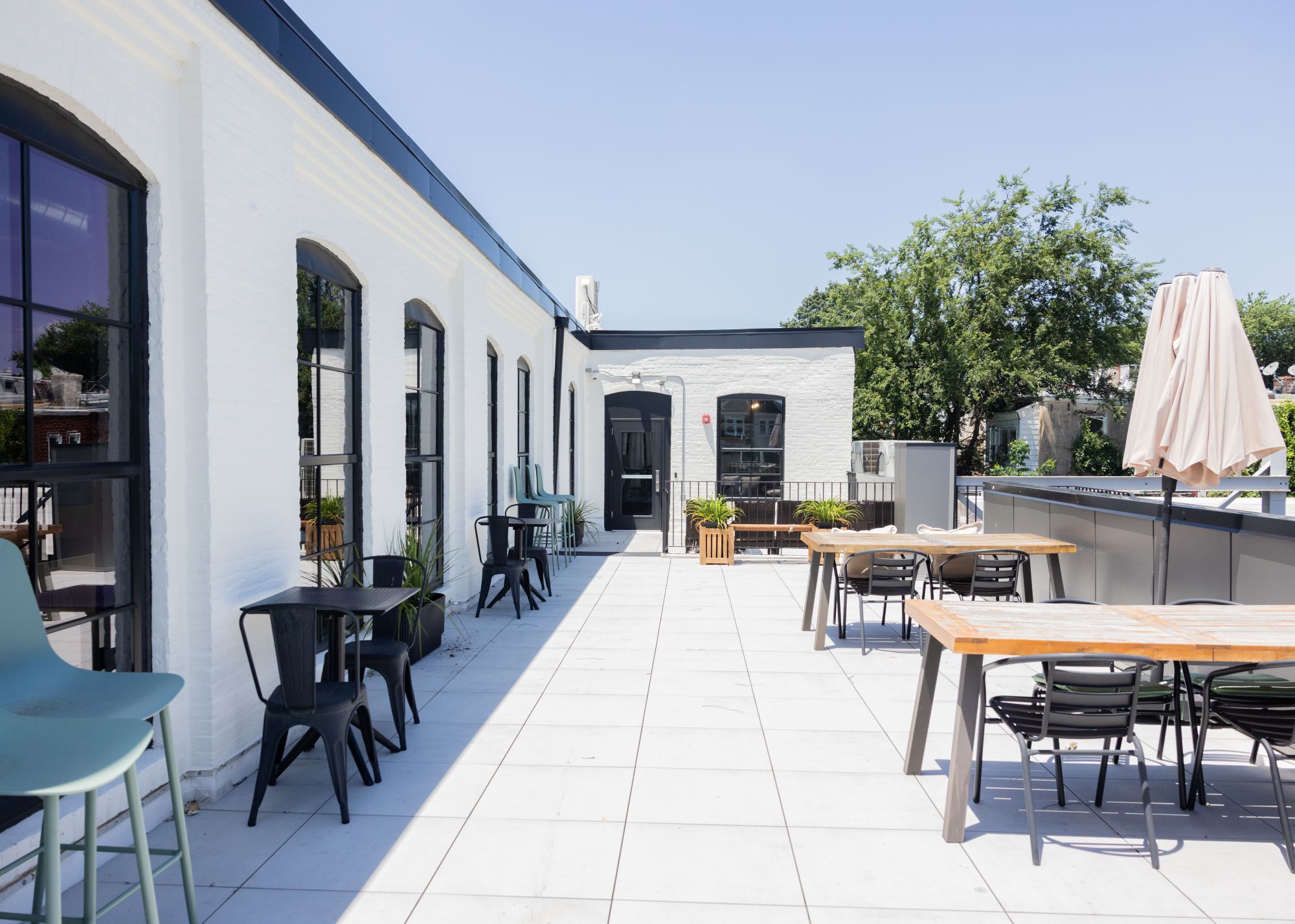 Outdoor patio with white tiled floor, black-framed windows, tables and chairs, potted plants, and an umbrella on a sunny day.