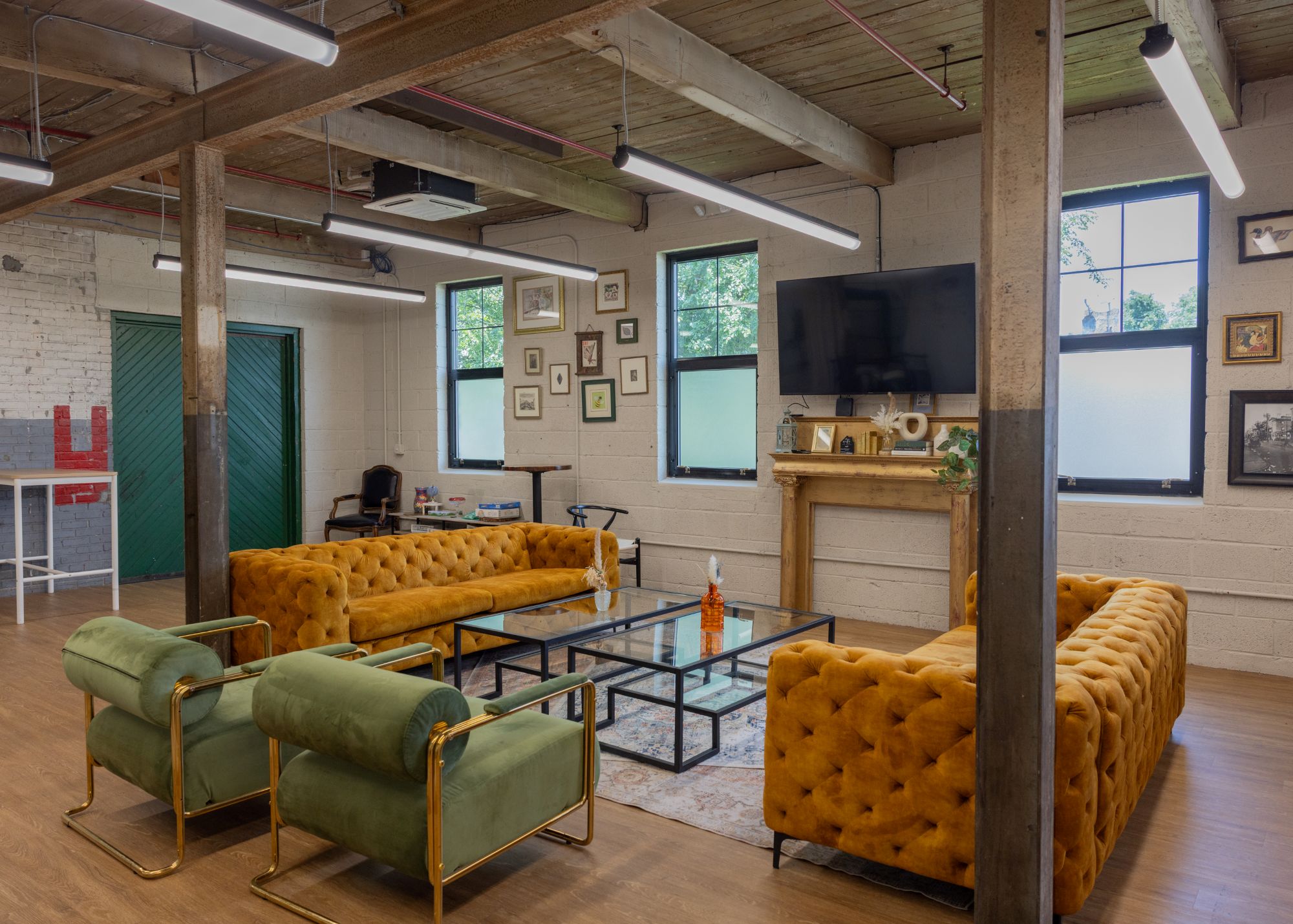 Modern office lounge with mustard yellow sofas, green chairs, glass coffee tables, wall decor, TV, and wooden floors under exposed beams and ceiling lights.