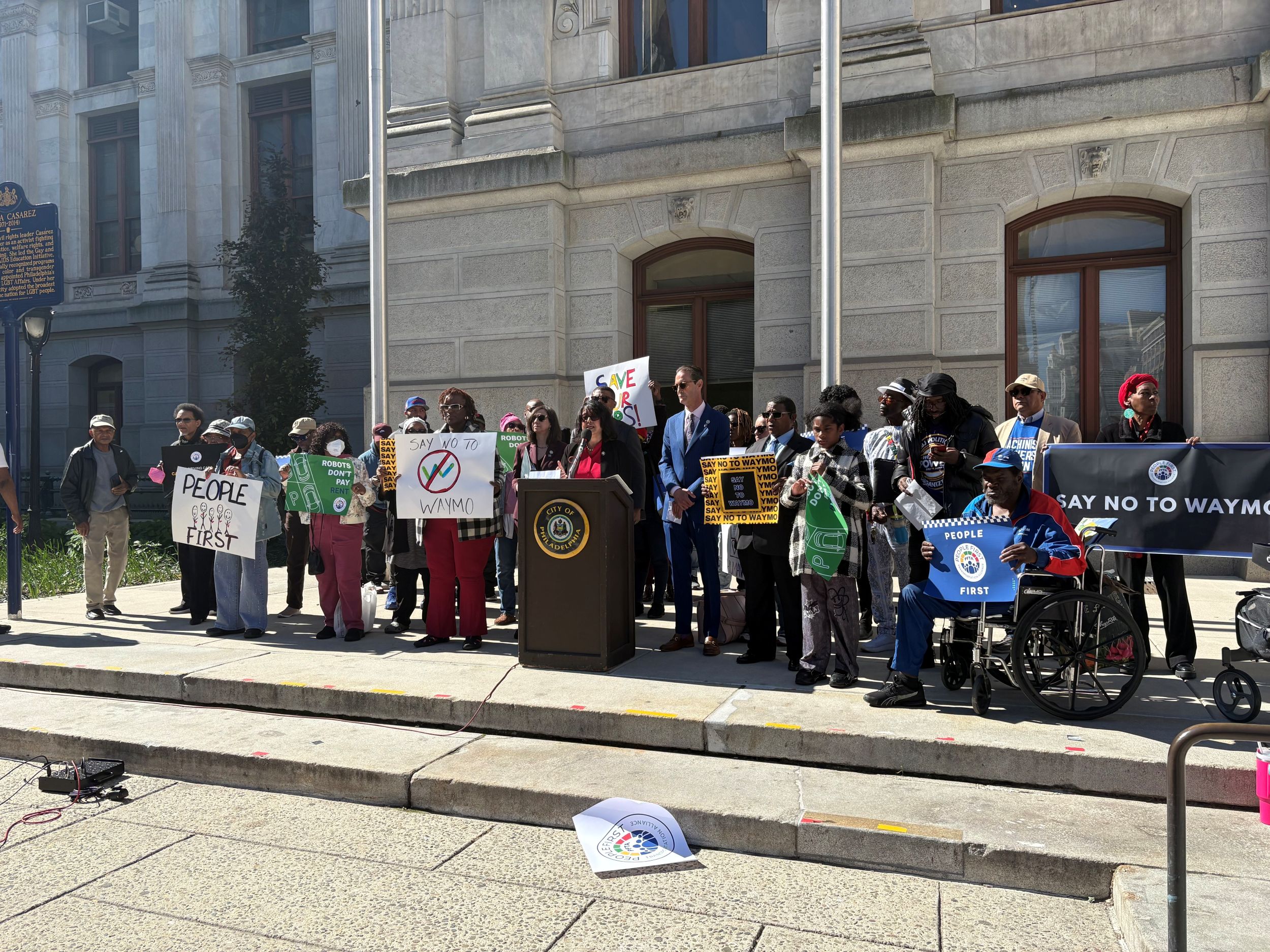 A group of people stand at a podium outside a government building, holding signs protesting and reading "Say No to Waymo" and "People First.