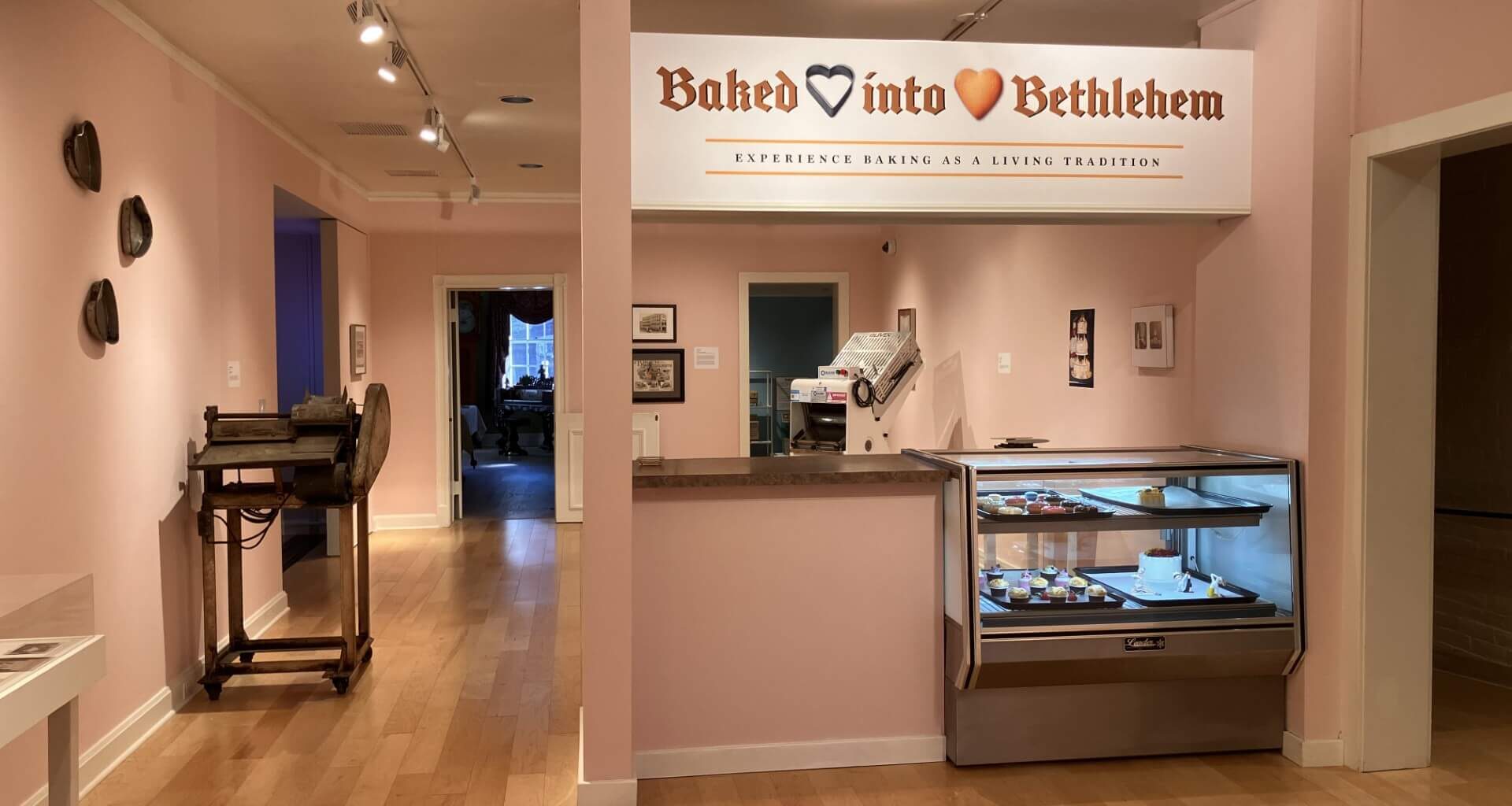 a museum gallery with pale pink walls and light wood floors, where a white sign reading "baked into bethlehem" hangs above a mock bakery counter with a glass display case of small cakes and pastries; an old dough-rolling machine sits on a cart against the wall to the left, with open doorways leading to other rooms