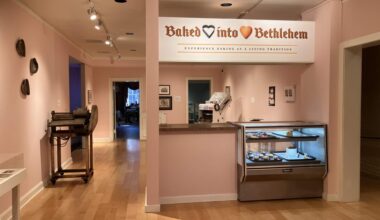 a museum gallery with pale pink walls and light wood floors, where a white sign reading "baked into bethlehem" hangs above a mock bakery counter with a glass display case of small cakes and pastries; an old dough-rolling machine sits on a cart against the wall to the left, with open doorways leading to other rooms