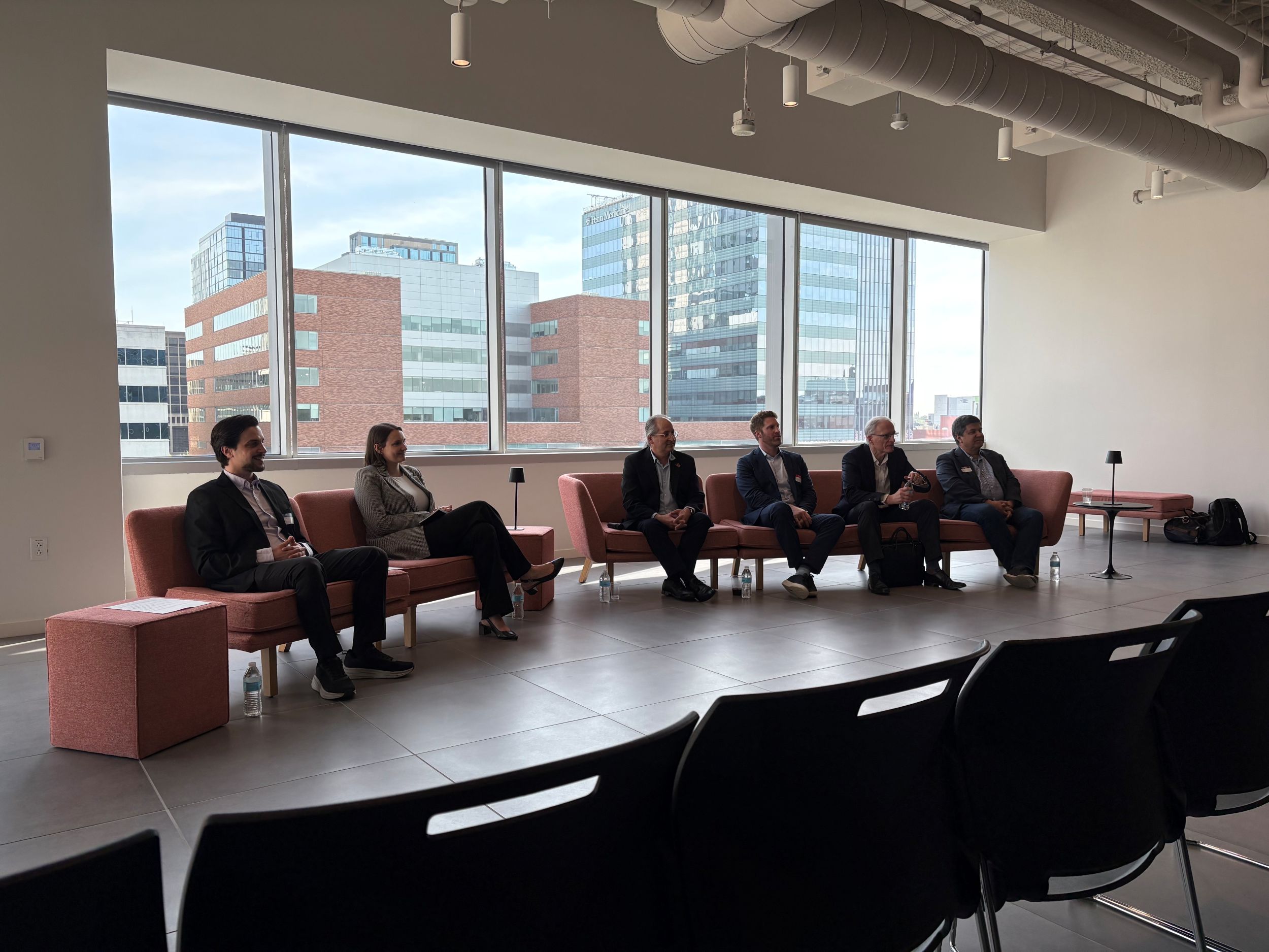 Six people in business attire sit on chairs in front of large windows with city buildings visible outside. Empty black chairs are in the foreground.