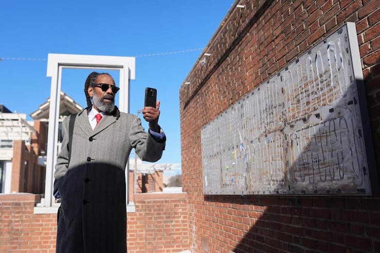 Man in overcoat and sunglasses holds up phone, with brick walls around him