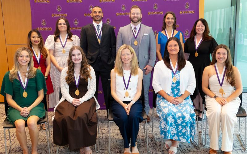 Group of award recipients wearing medals pose for a photo in front of a University of Scranton backdrop.