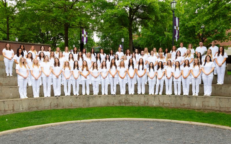 Large group of students in matching white uniforms posed on tiered stone steps outdoors, with trees and campus banners in the background.