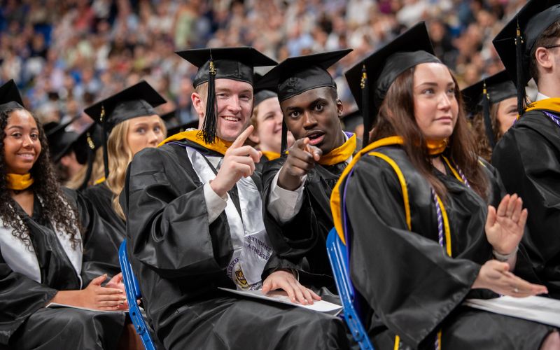 Graduates in caps and gowns seated at a commencement ceremony, with two students in the center pointing toward the camera.