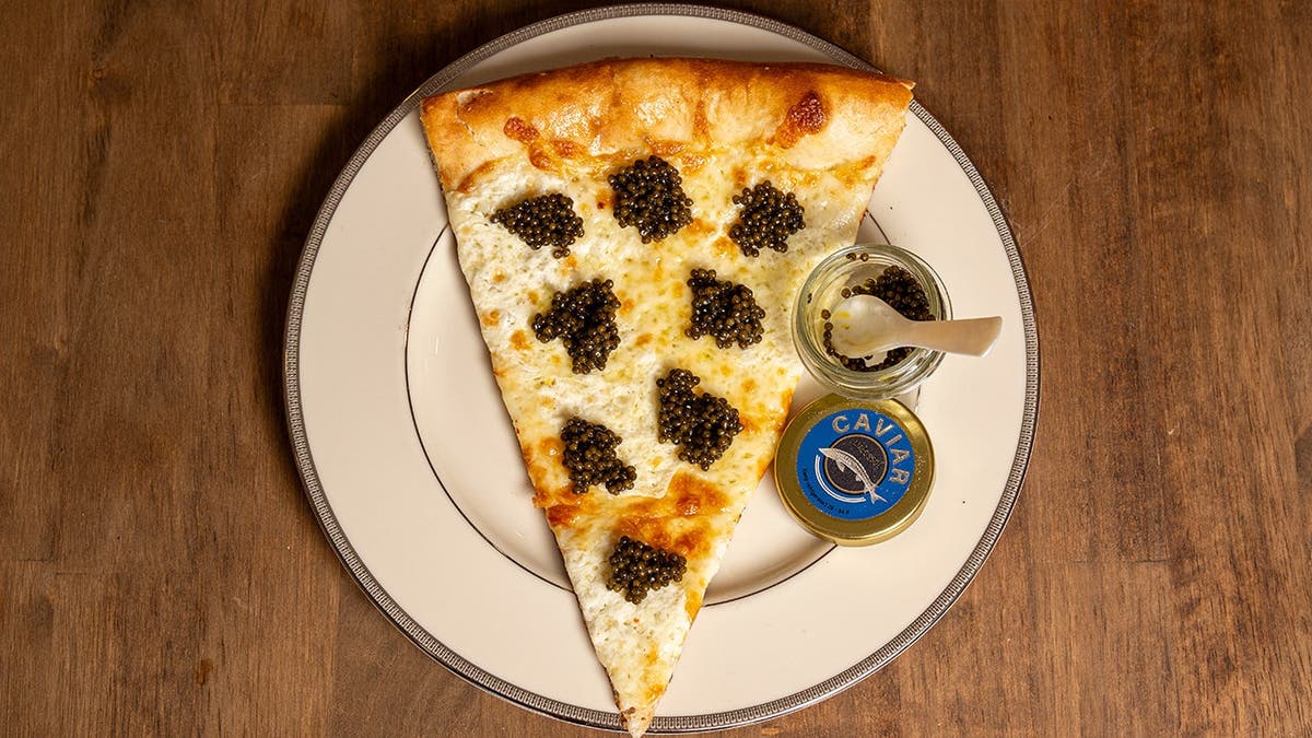 A piece of white pizza sits on a white plate, covered in caviar with a jar of caviar with a spoon beside it.