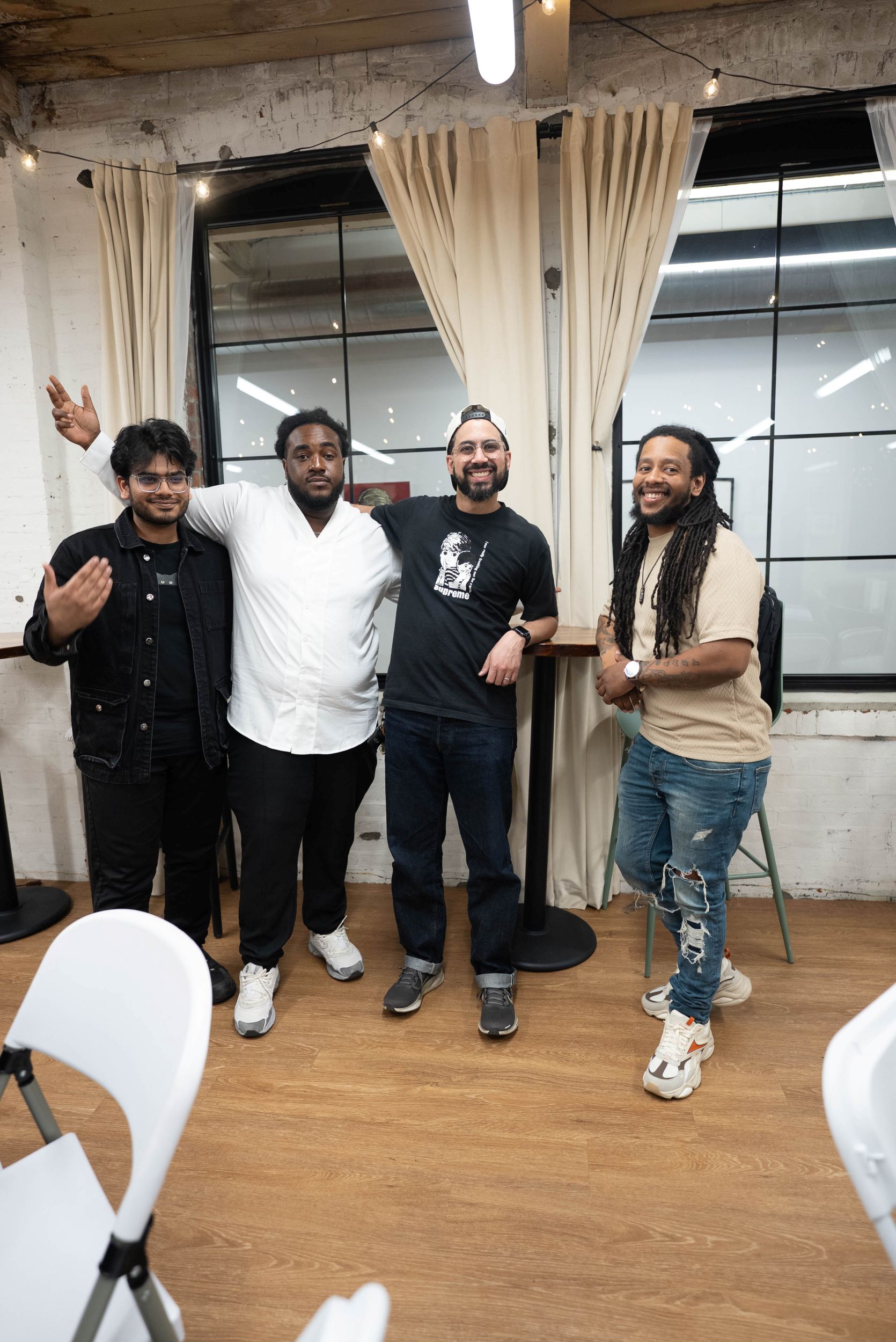 Four men stand together and pose for a photo in a casual indoor setting with large windows and string lights.