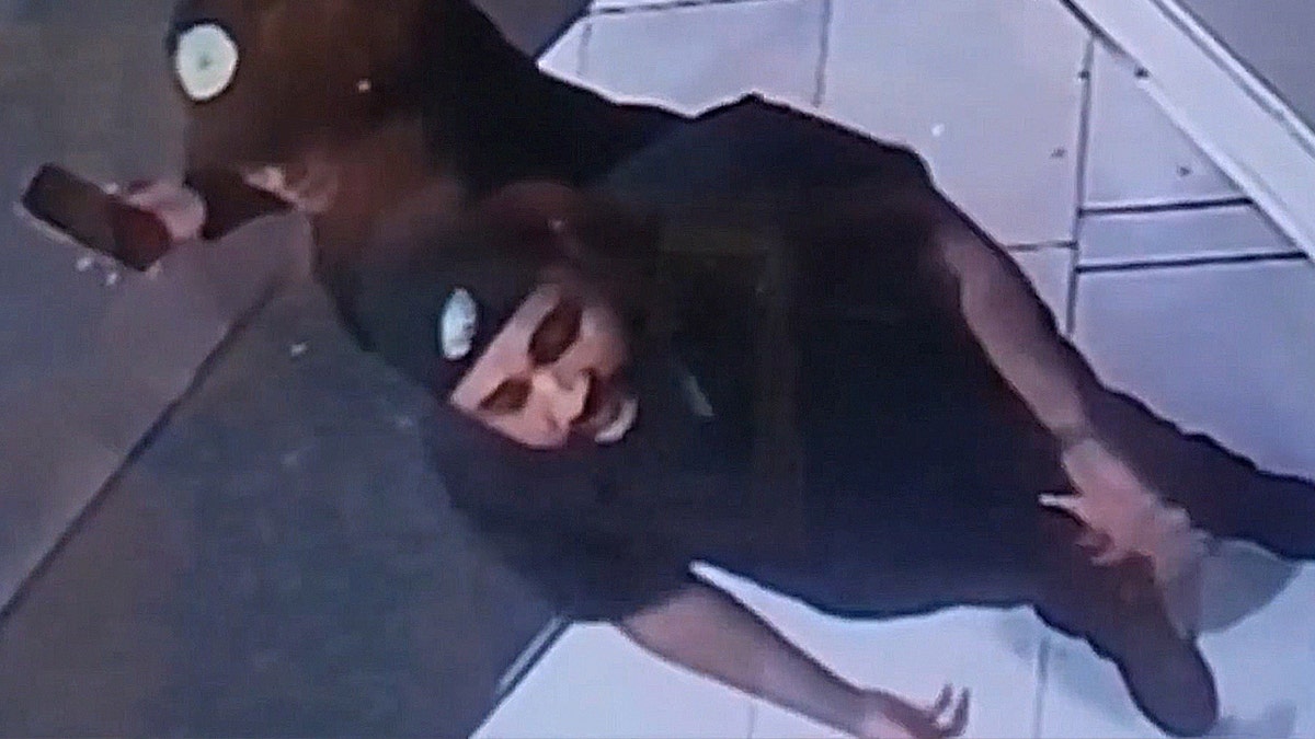 A grainy, high-angle surveillance still captures the face of a man wearing a dark beanie and dark clothing.
