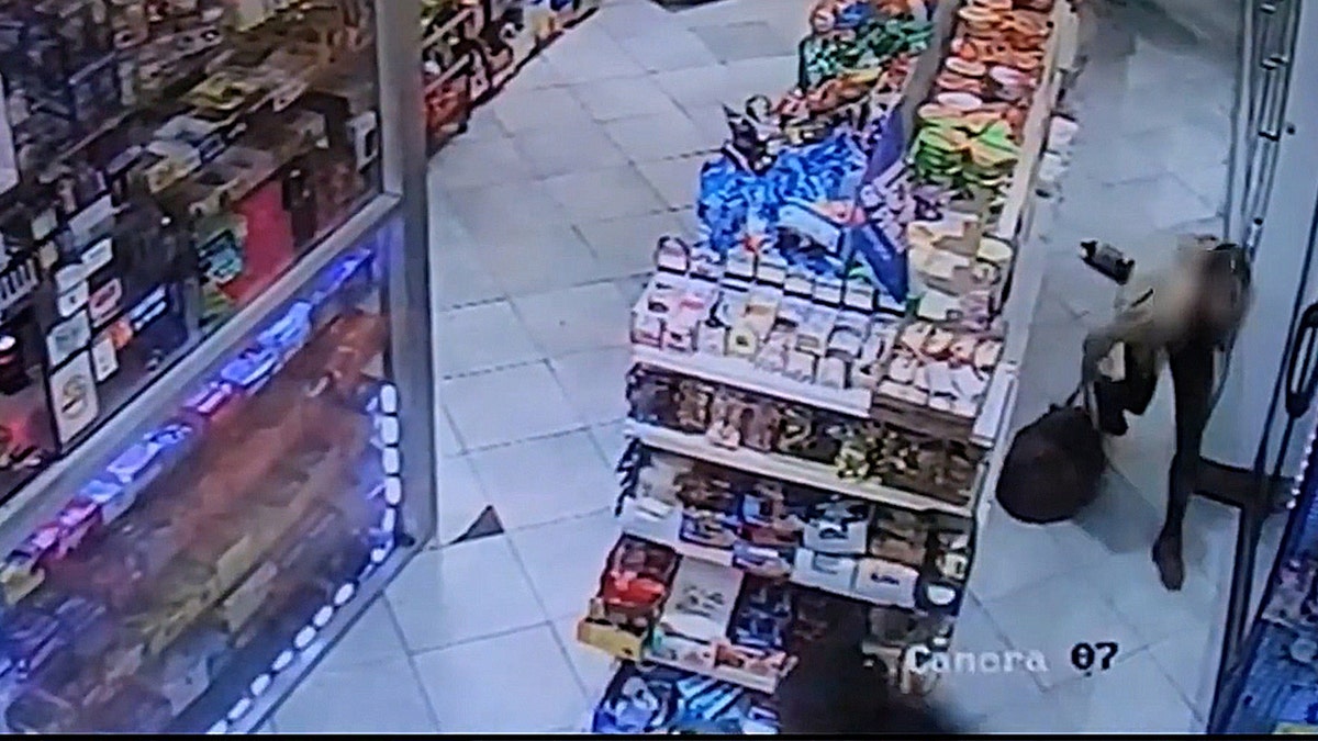 Security camera view of a store aisle with shelves of chips and snacks while a person moves toward the exit.