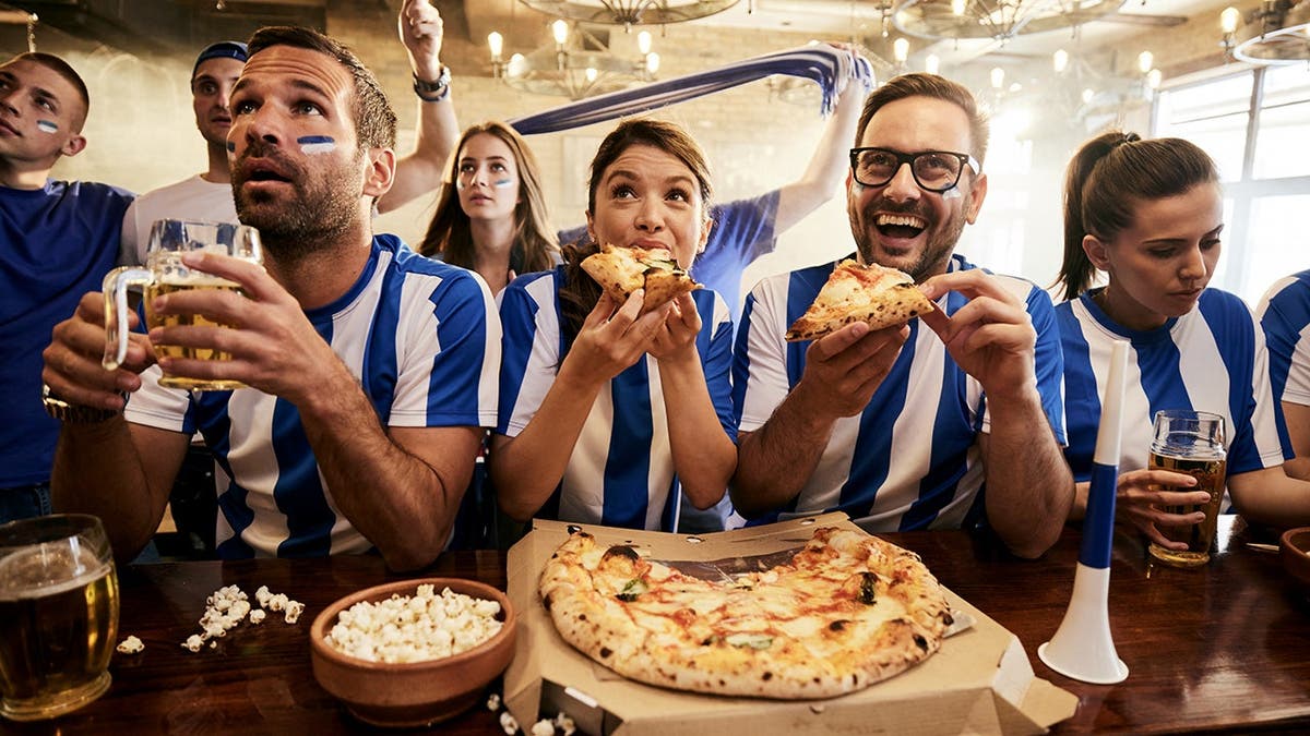 A group of sports fans in jerseys eats pizza while watching a game.