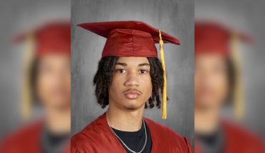 Tymell Millan-Mason poses for his Lincoln Leadership Academy Class of 2026 senior portrait, wearing a red graduation cap and gown with a gold tassel over a black t-shirt and silver chain.