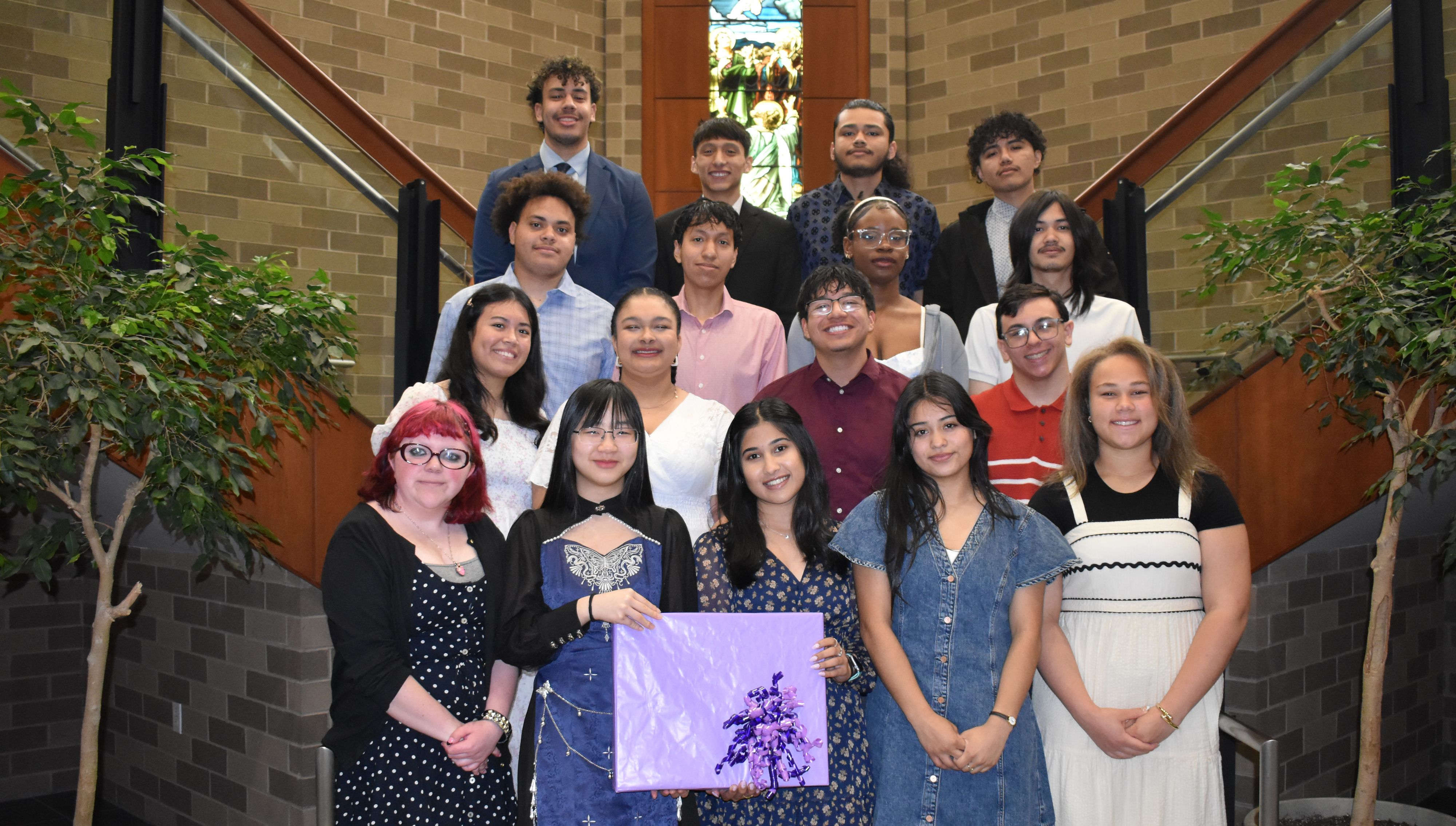 Group of students pose on an indoor staircase in a brick building, with stained-glass windows behind them and one person holding a purple gift bag.