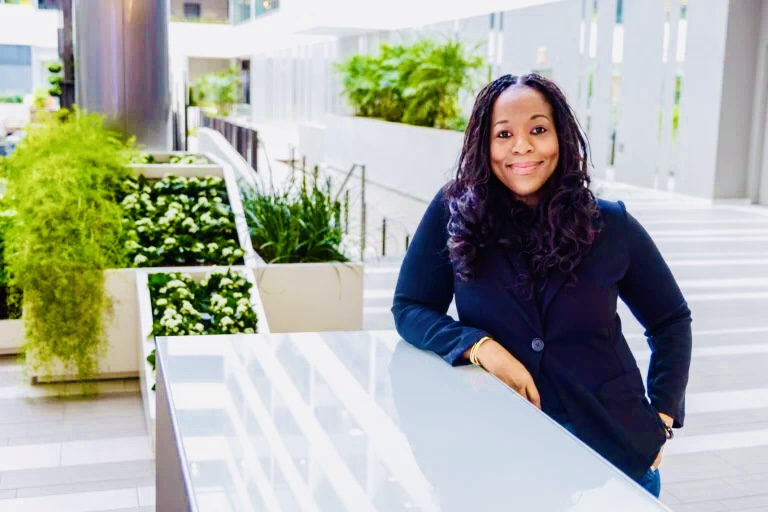 A woman in a dark blazer stands by a reflective table in a modern indoor space with plants and natural light.
