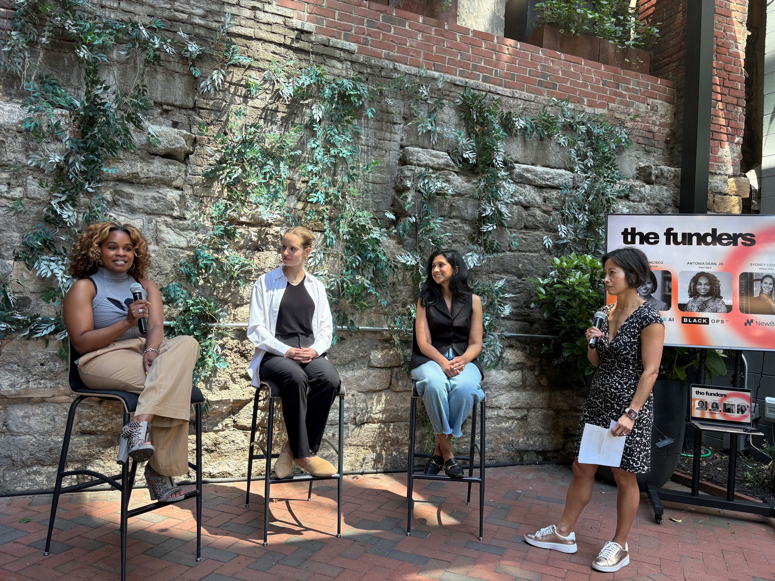 Four women participate in an outdoor panel discussion in front of a stone wall decorated with greenery; one woman stands holding a microphone and notes, while the others sit on stools.