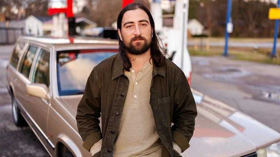 Noah Kahan poses by brown wagon