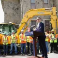 The governor stands at a podium outside, with construction workers in yellow and orange vests stand in front of large yellow construction equipment.