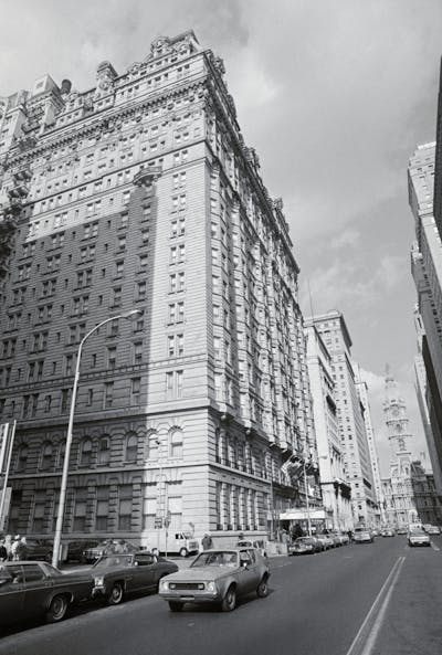 Black-and-white photo of large ornate building alongside two-way street with 1970s-era cars