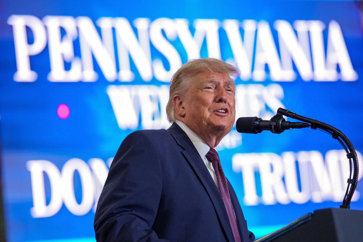An individual, dressed formally in a suit and tie, is delivering a speech at a podium equipped with a microphone. The backdrop features a large sign with the text "PENNSYLVANIA" prominently displayed, along with additional words that are partially obscured.