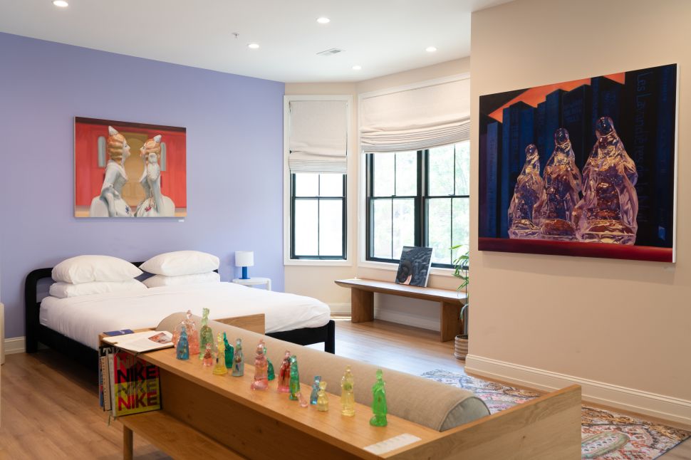 An art-filled bedroom shows a white bed, pastel walls, large figurative paintings, colorful small sculptures lined on a wooden bench and daylight coming through tall windows.