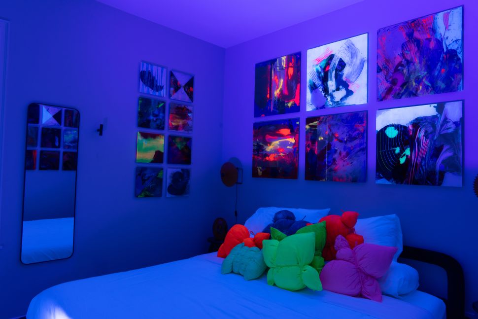 An installation-style bedroom is lit in blue-purple light, with neon-colored abstract paintings on the walls and bright sculptural pillows piled on a white bed.