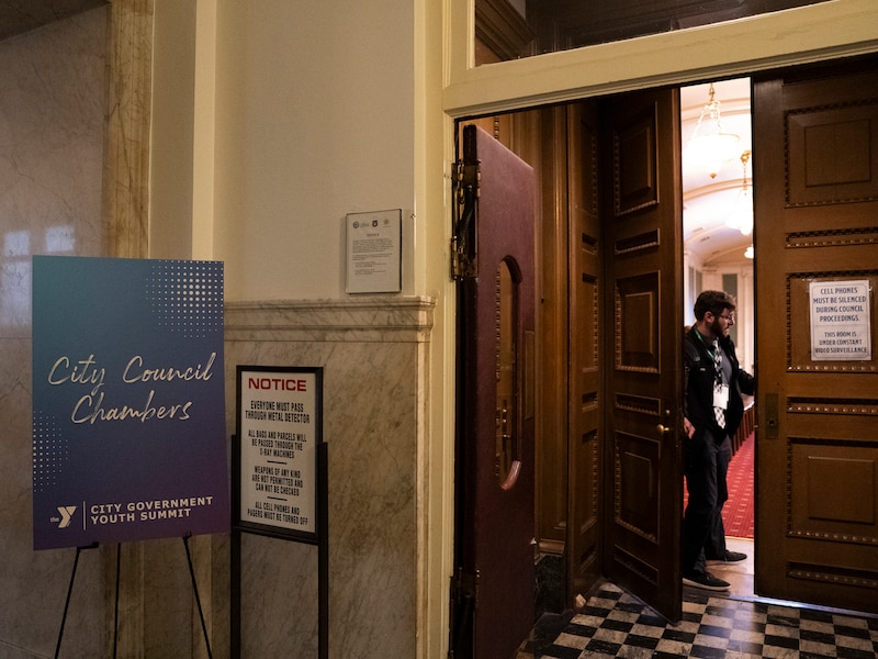 A photograph of a display board reads "City Council Chambers," standing on the left side, with a white man closing the chamber's door on the right.