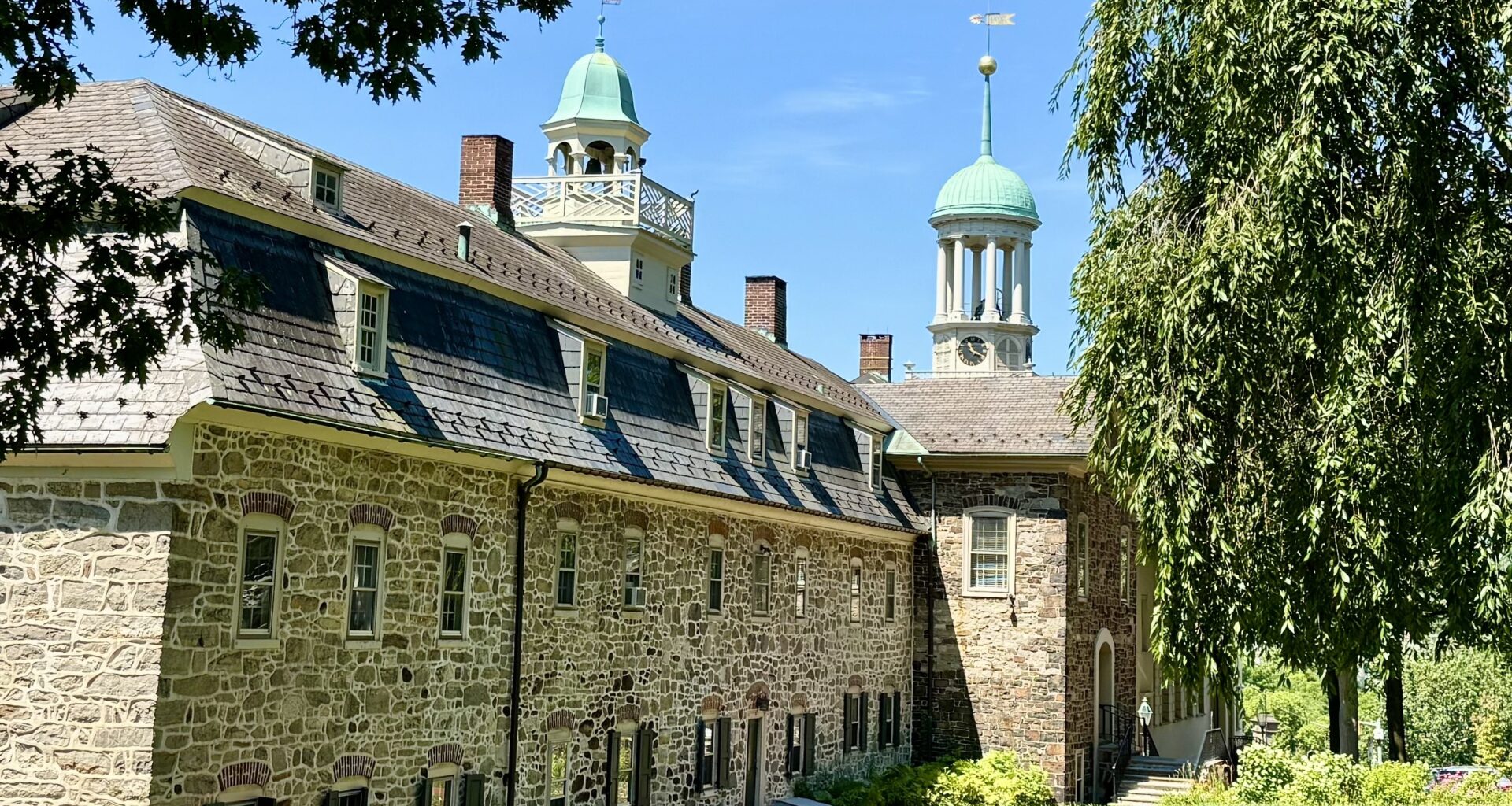 To the left, a stone building stands with multiple windows and a belfry; on the right is a large tree.