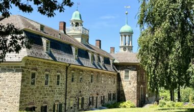 To the left, a stone building stands with multiple windows and a belfry; on the right is a large tree.