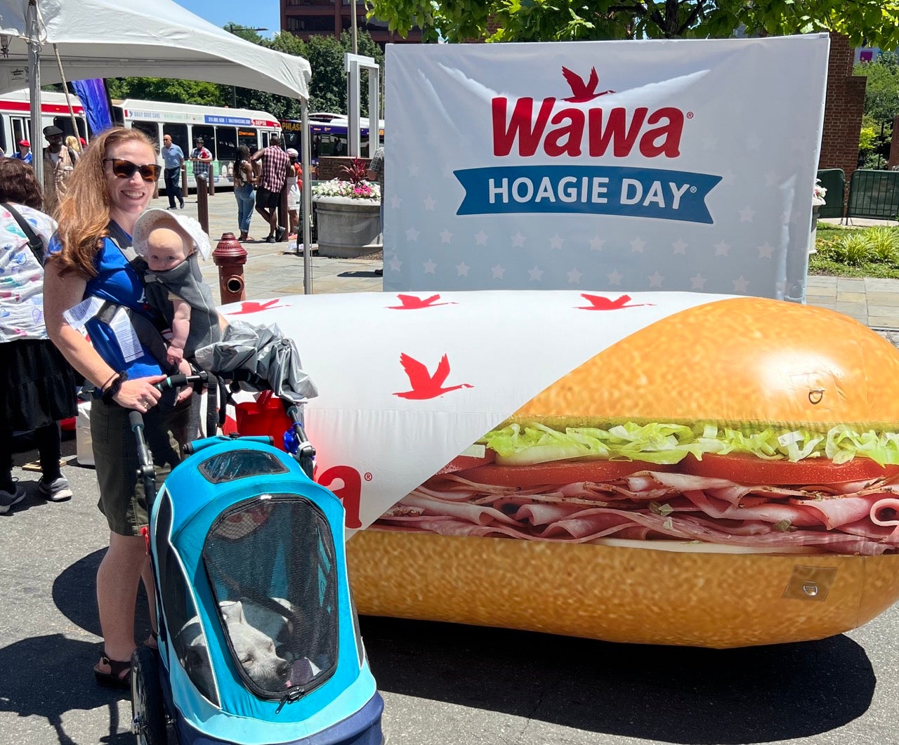 Baby's first WAWA Hoagie Day. Kristen wears baby Trudy in a carrier in front of her, posing in front of a WAWA Hoagie Day sign with a giant inflatable hoagie and her dog Gloria in a blue stroller in front of her.