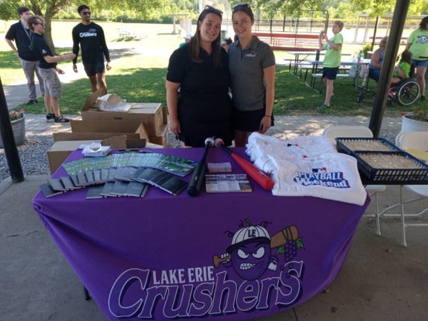 Regan Cole, left, account manager for the Lake Erie Crushers, and Ruby Wallace, also with the Crushers, at the merchandise table during Play Ball Weekend at Volunteer Field, which is located on the border of Sheffield Village and Sheffield Lake. (Bob Stascak - For The Morning Journal)