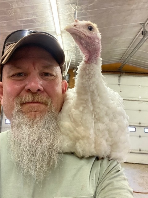 A man with a white beard poses for a selfie-style photo with a turkey or other bird sitting on his shoulder