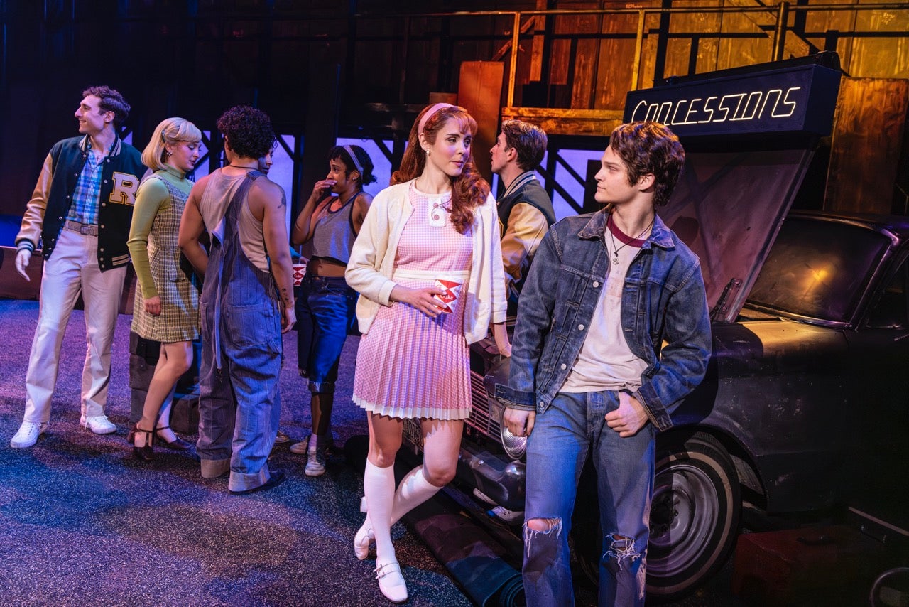 (L-R) Emma Hearn dressed as a soc (preppy socialite) and Nolan White (dressed as a greaser, jeans and a jean jacket) lean on a car talking among a bunch of other teens on stage in The Outsiders North American Tour