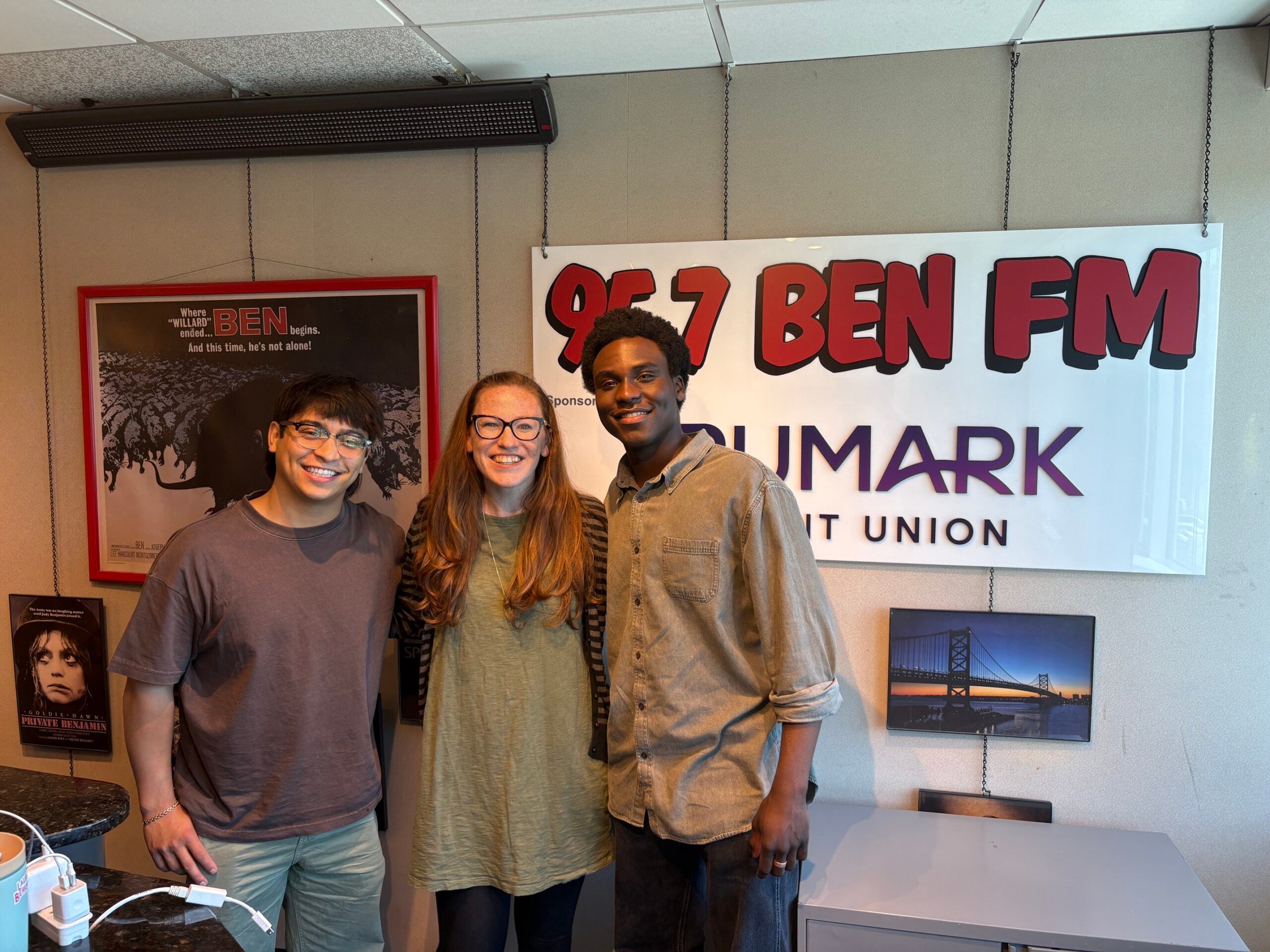 Tyler Jordan Wesley and Bonale Fambrini from the Outsiders National Touring Cast pose with Kristen Herrmann from BEN FM (middle) in front of the white BEN FM studio sponsor sign that reads 95.7 BEN FM - Trumark Credit Union in red and purple letter.