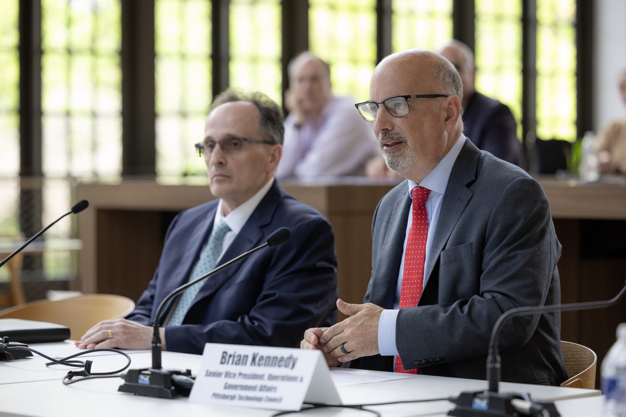 Two men in suits sit at a conference table with microphones; one is speaking while the other listens. Nameplates and people are visible in the background.