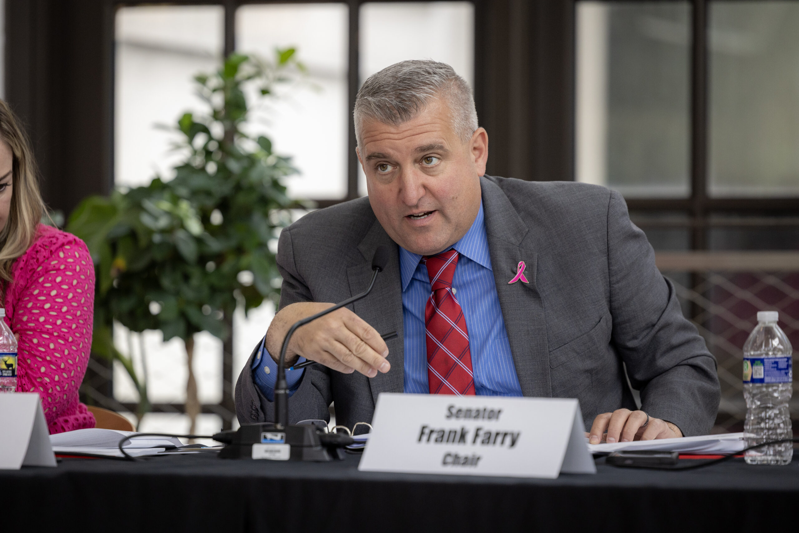 A man in a suit and tie, wearing a pink ribbon, sits at a table with a nameplate that reads "Senator Frank Farry Chair" and gestures while speaking into a microphone.